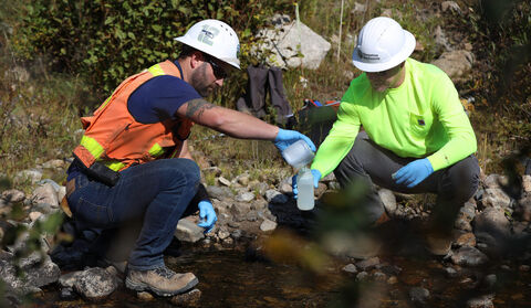 Two men in safety gear collect water samples from a stream.