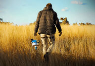 A worker walks through tall, dry grass carrying an electronic device.