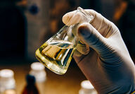 A gloved hand holds a beaker of gold-colored liquid in a lab.