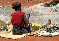 African woman sitting on ground using sledgehammer to break rocks.